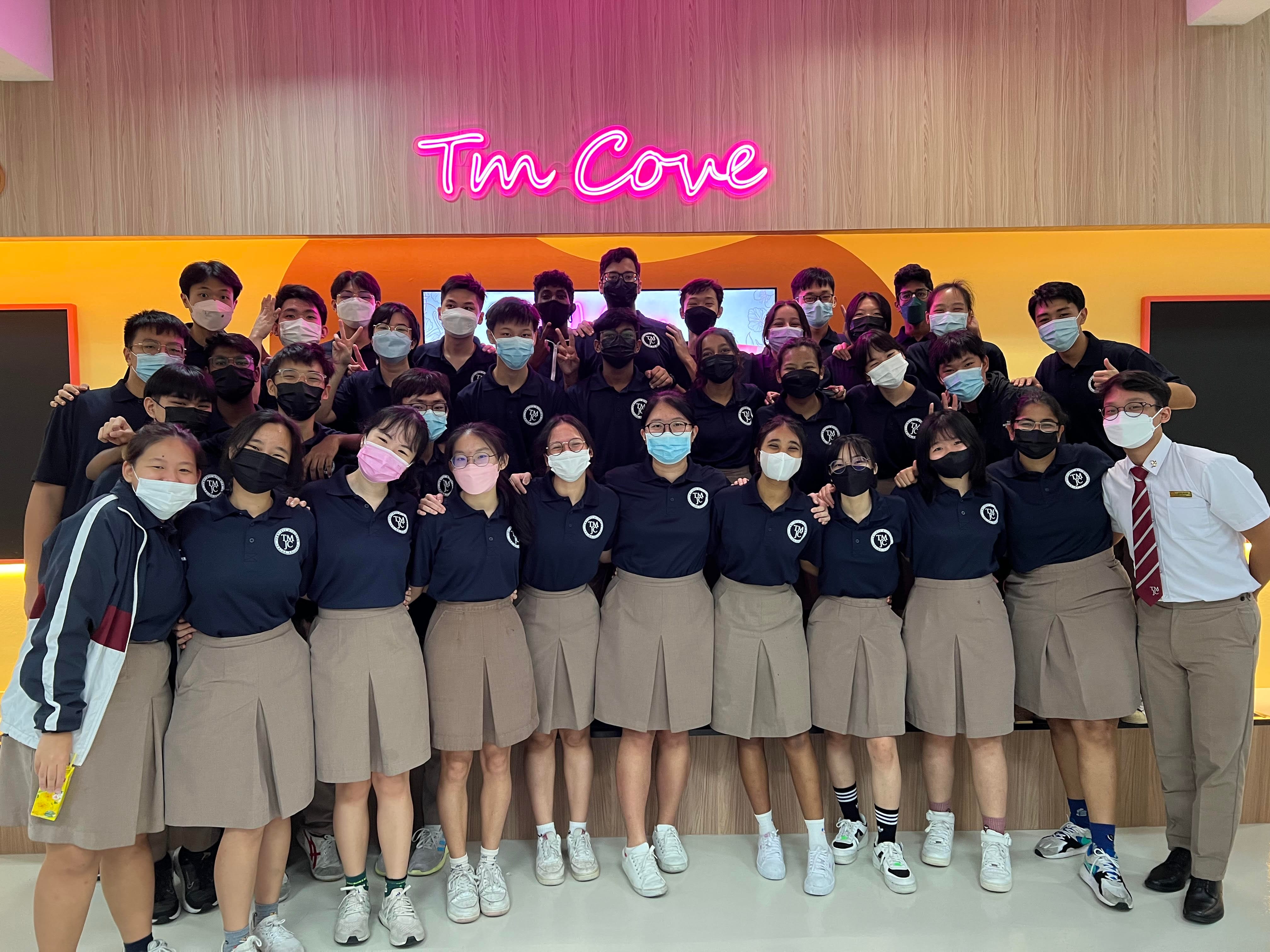 Large group of masked students in navy uniforms posing under a pink neon sign reading "Tm Cove."